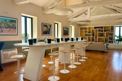 Salle lumineuse avec un long comptoir blanc, des tabourets, des bouteilles exposées et des poutres apparentes au plafond.