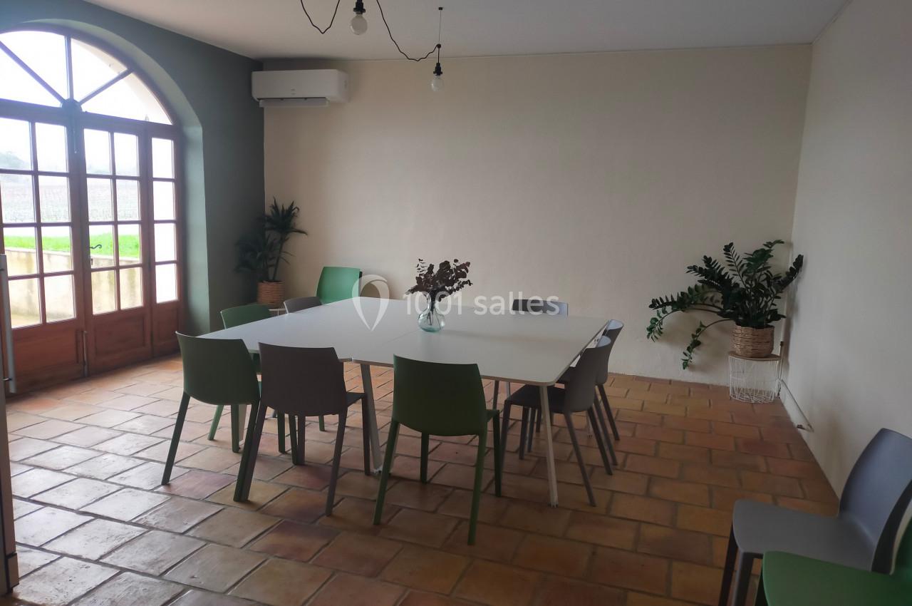 Salle lumineuse avec table blanche entourée de chaises colorées, plantes décoratives et grande porte vitrée.