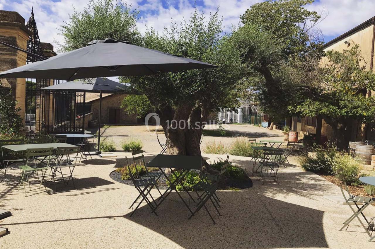 Terrasse extérieure avec tables, chaises et parasols, entourée d'arbres et de végétation sous un ciel partiellement nuageux.