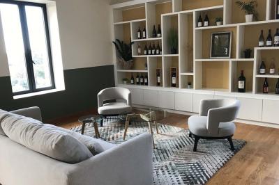 Façade d'un bâtiment clair avec une terrasse aménagée, un grand arbre et un ciel bleu en arrière-plan.