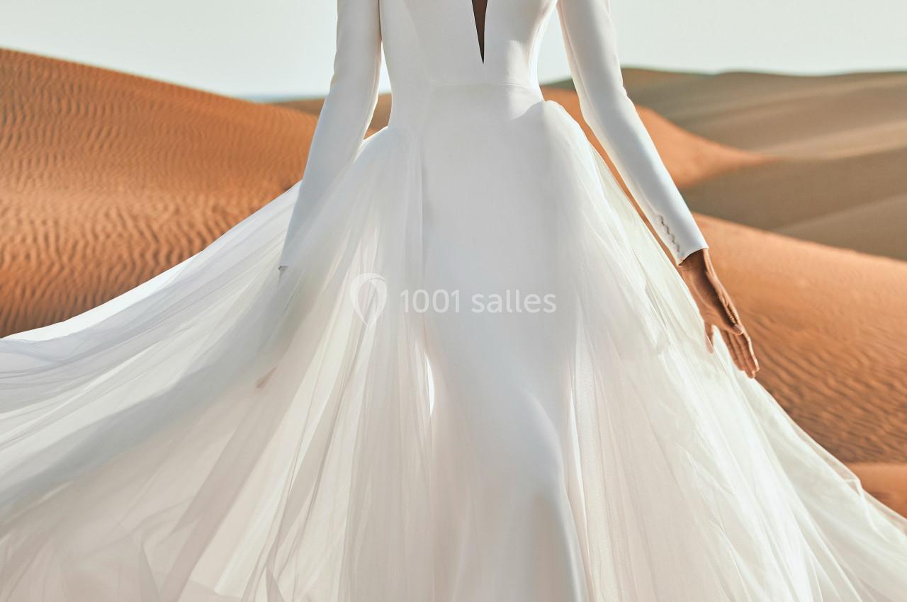 Femme portant une robe de mariée blanche fluide avec manches longues, posant dans un désert sous un ciel clair.