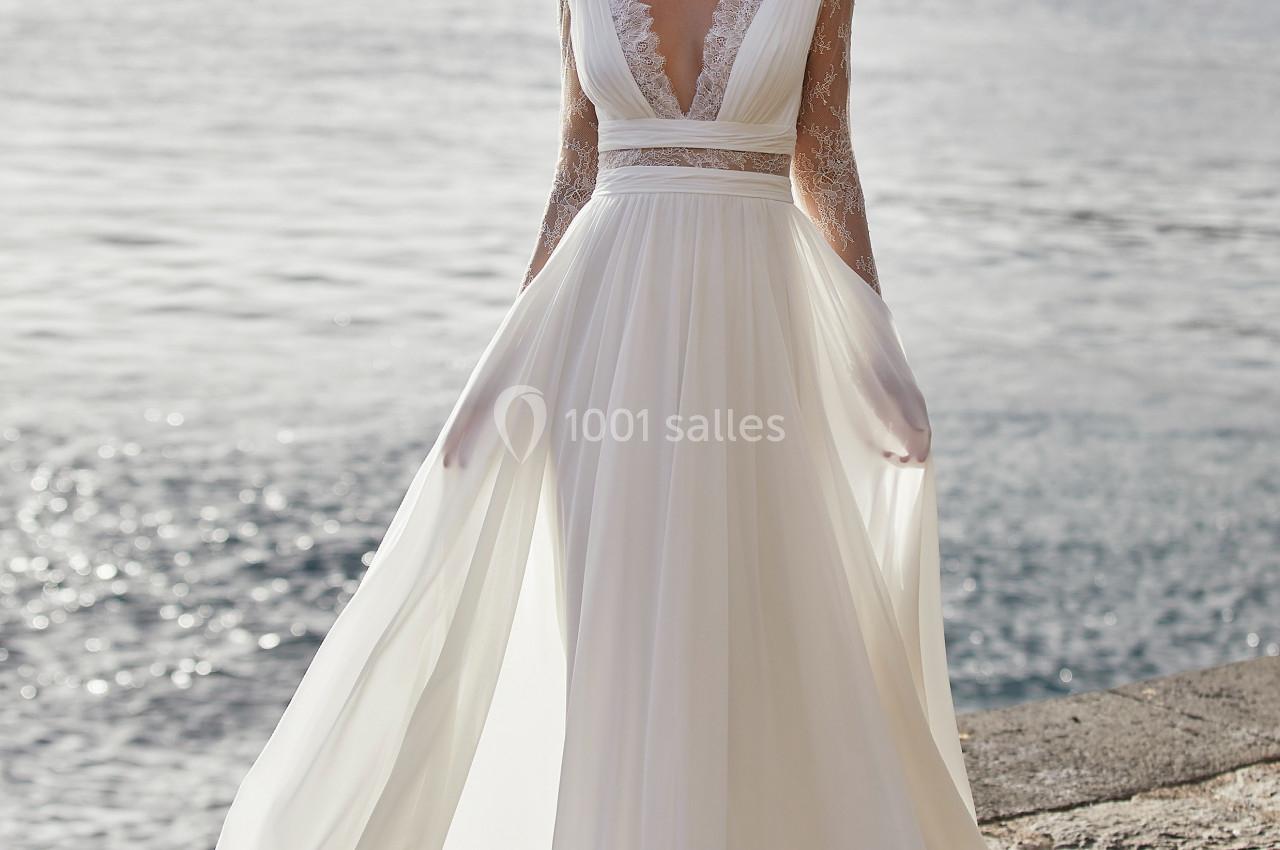 Femme portant une robe de mariée fluide avec dentelle, debout près d'une étendue d'eau et de rochers.