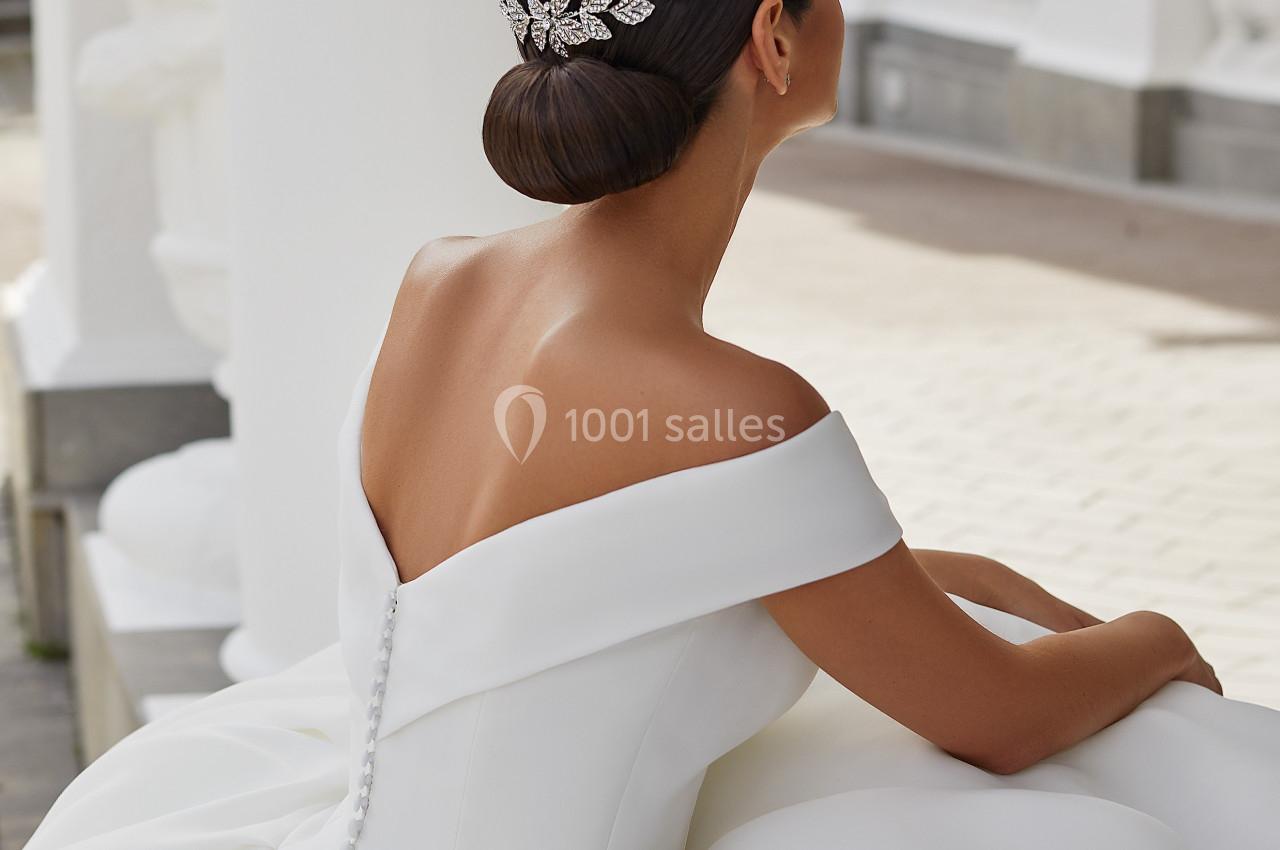 Femme en robe de mariée blanche avec un chignon orné d'un accessoire argenté, assise près d'une architecture classique.
