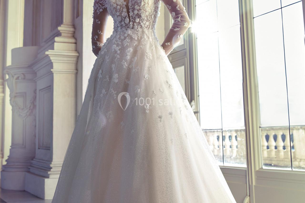 Femme portant une robe de mariée élégante avec manches longues en dentelle, dans une pièce lumineuse avec de grandes…