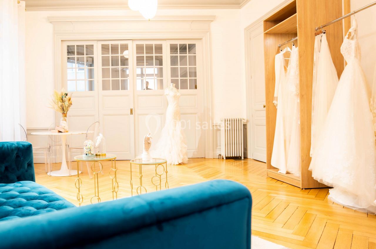 Salon lumineux avec canapé bleu, robes de mariée suspendues et mannequin exposant une robe dans un décor élégant.