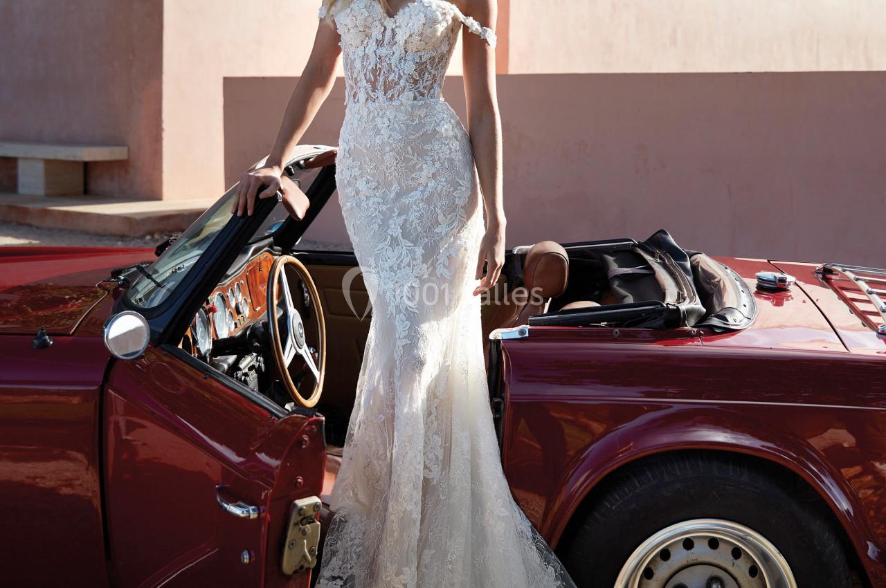 Femme en robe de mariée en dentelle, debout près d'une voiture décapotable rouge dans un décor extérieur ensoleillé.