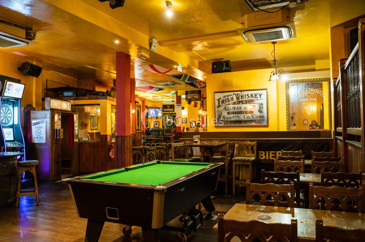 Salle de bar chaleureuse avec un billard au centre, des tables en bois et des décorations murales variées.
