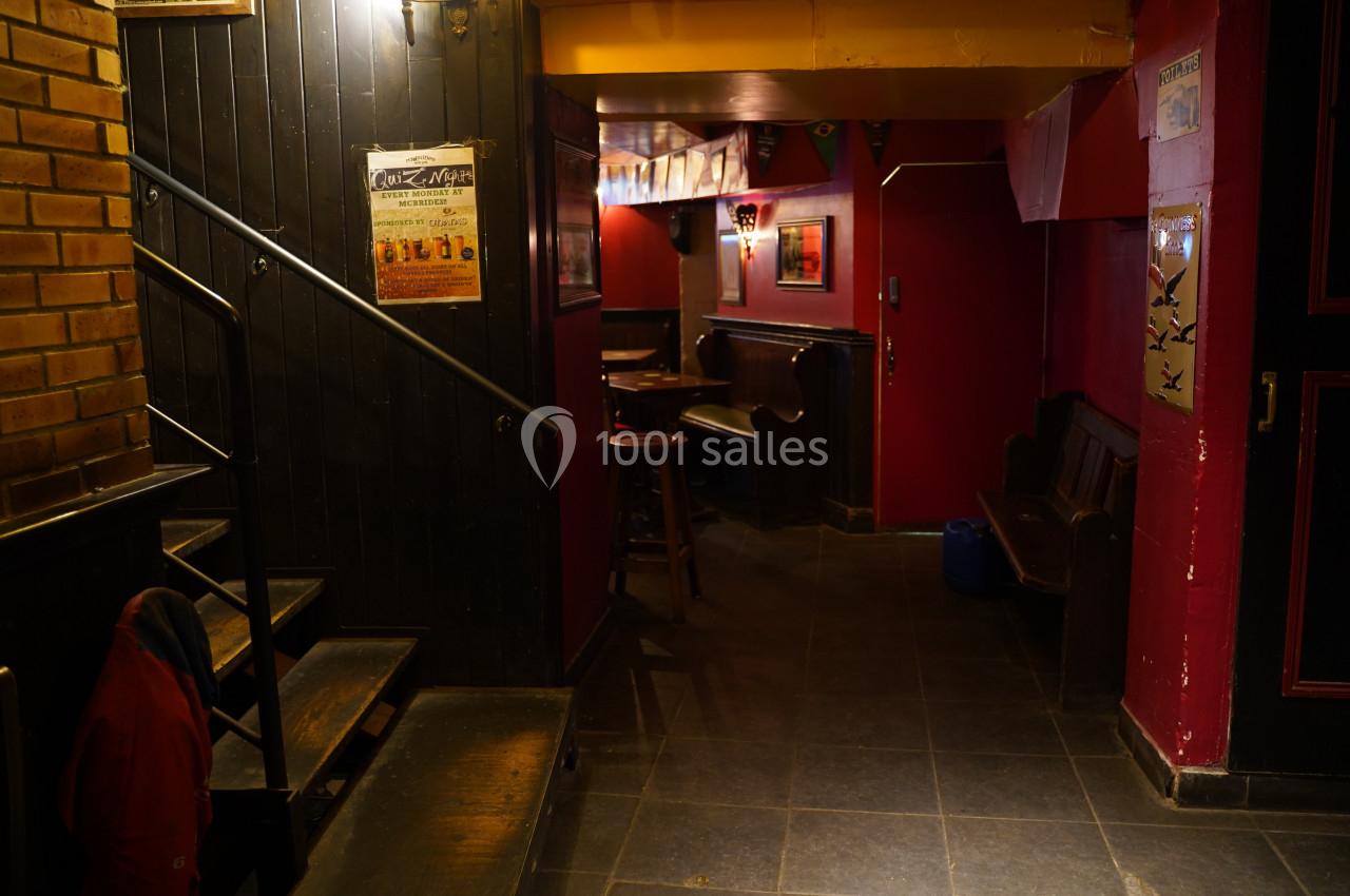 Couloir sombre avec murs rouges et noirs, affiches accrochées, escalier à gauche et mobilier de bar en bois.