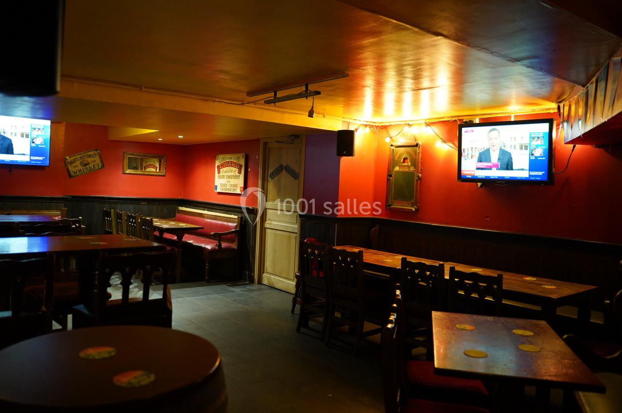 Salle de bar avec tables en bois, banquettes rouges, murs rouges et deux télévisions diffusant des informations.