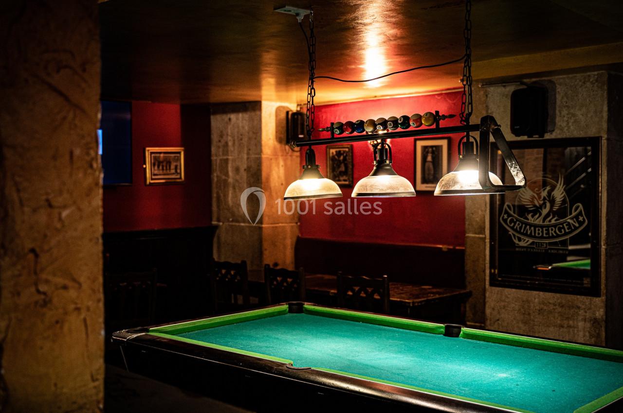 Table de billard éclairée par des suspensions dans un bar à l'ambiance tamisée avec murs rouges et décorations murales.