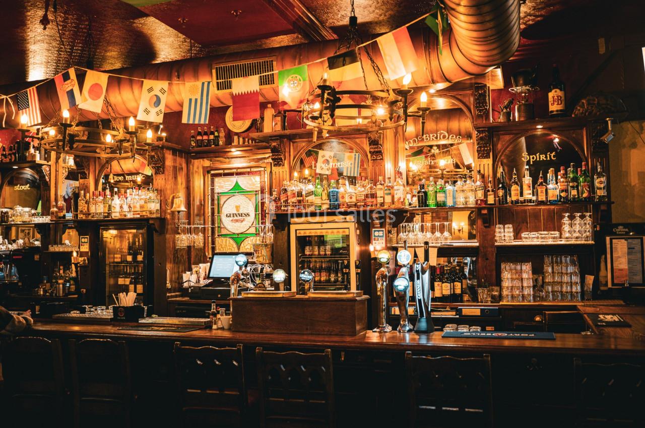 Intérieur d'un pub avec un bar en bois, des bouteilles alignées, des drapeaux suspendus et une ambiance chaleureuse.