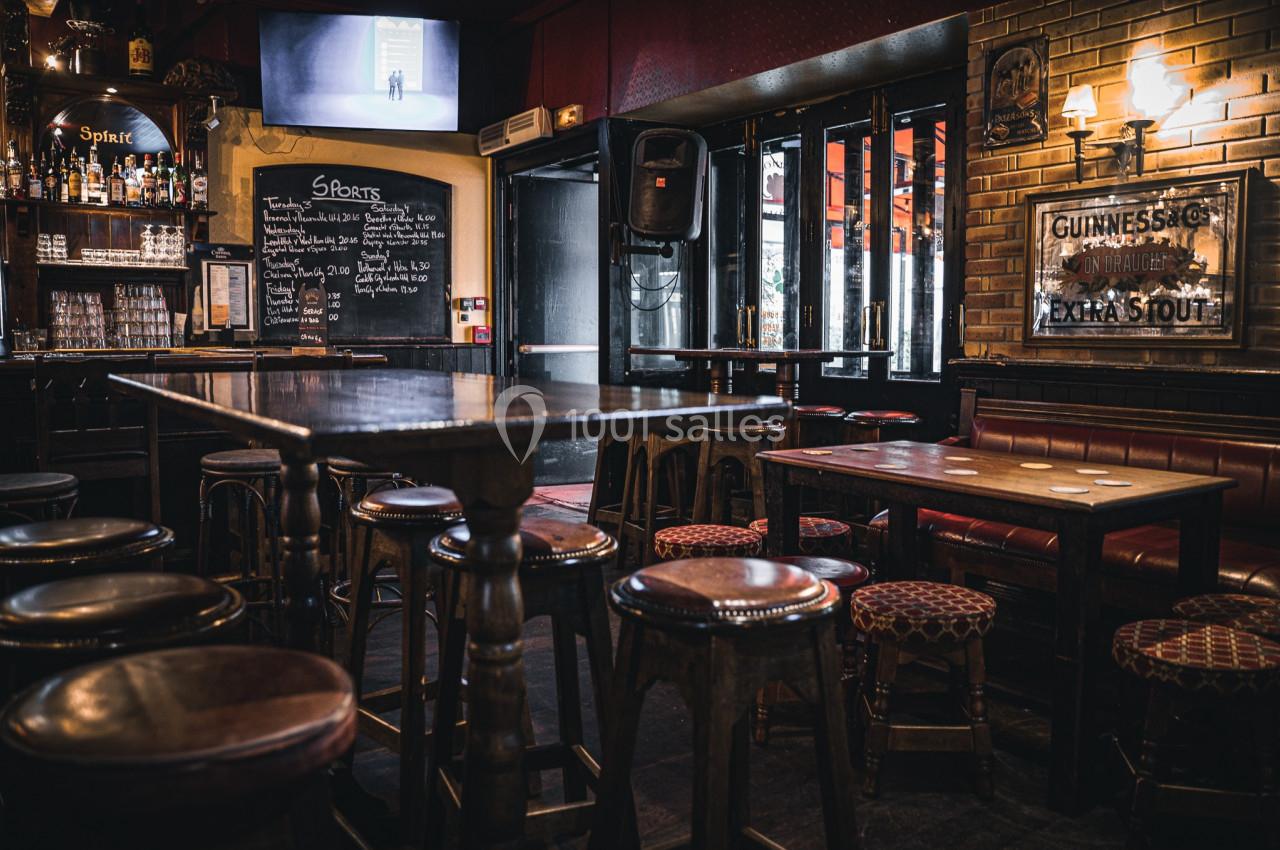 Intérieur d'un pub avec des tables en bois, des tabourets, un tableau noir et une ambiance tamisée.