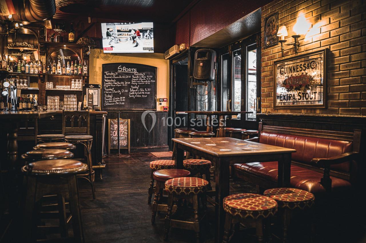 Intérieur d'un pub avec bar, tabourets, tables en bois, éclairage chaleureux et écran diffusant une émission sportive.