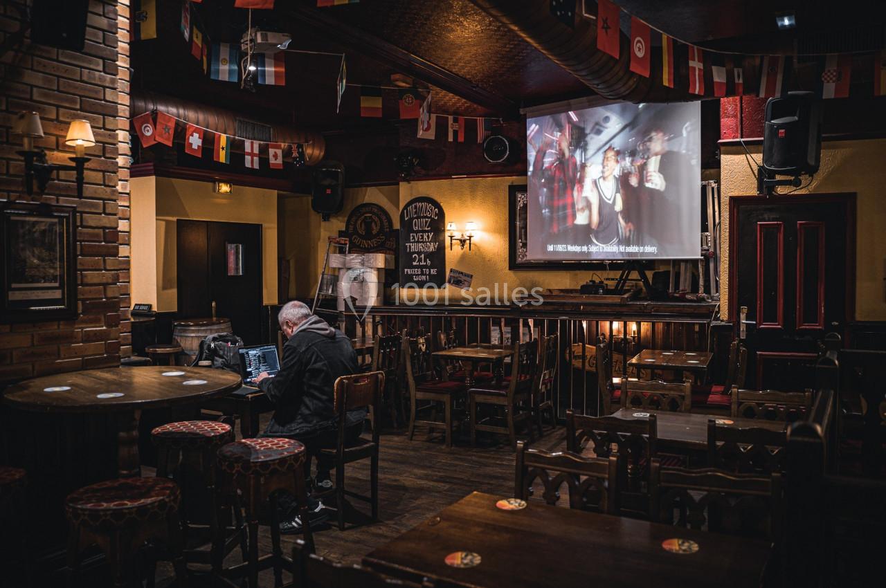 Un homme assis seul dans un pub vide, utilisant un ordinateur portable, avec un écran projetant une image en arrière-plan.