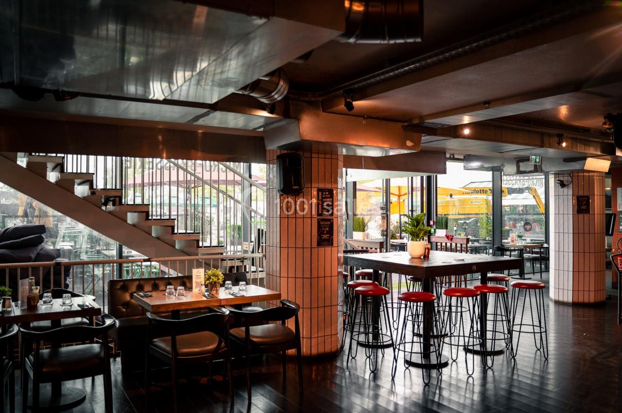 Intérieur d'un restaurant moderne avec tables en bois, tabourets rouges et grande baie vitrée donnant sur une terrasse.