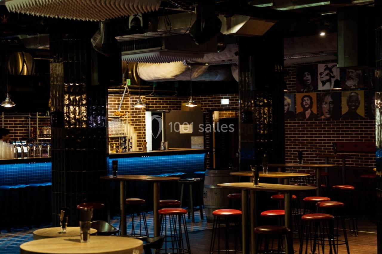 Salle de bar avec tables hautes, tabourets rouges et éclairage tamisé, décor industriel en arrière-plan.