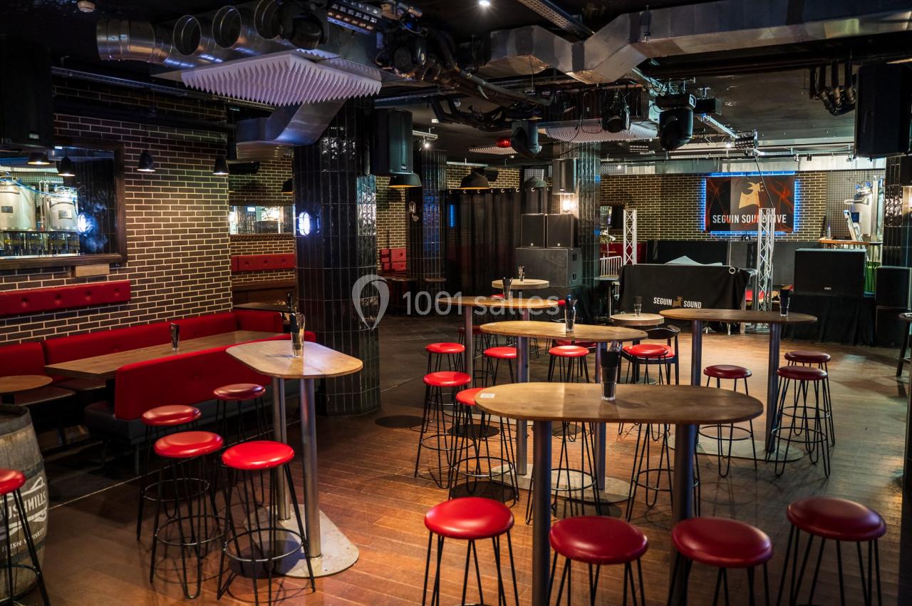 Salle de bar avec tables hautes, tabourets rouges, banquettes et éclairage tamisé dans un décor industriel.