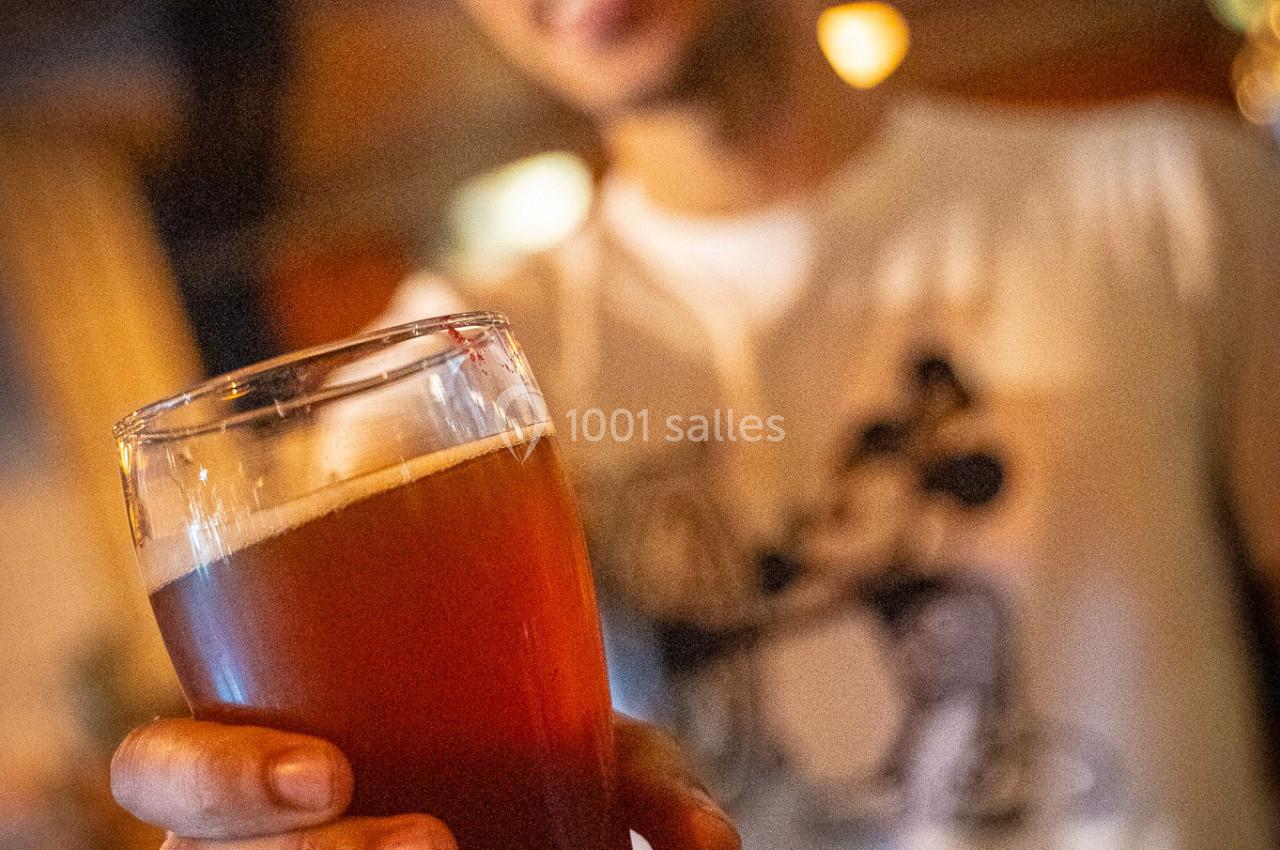 Un homme tend un verre de bière ambrée dans un environnement de bar ou de café.