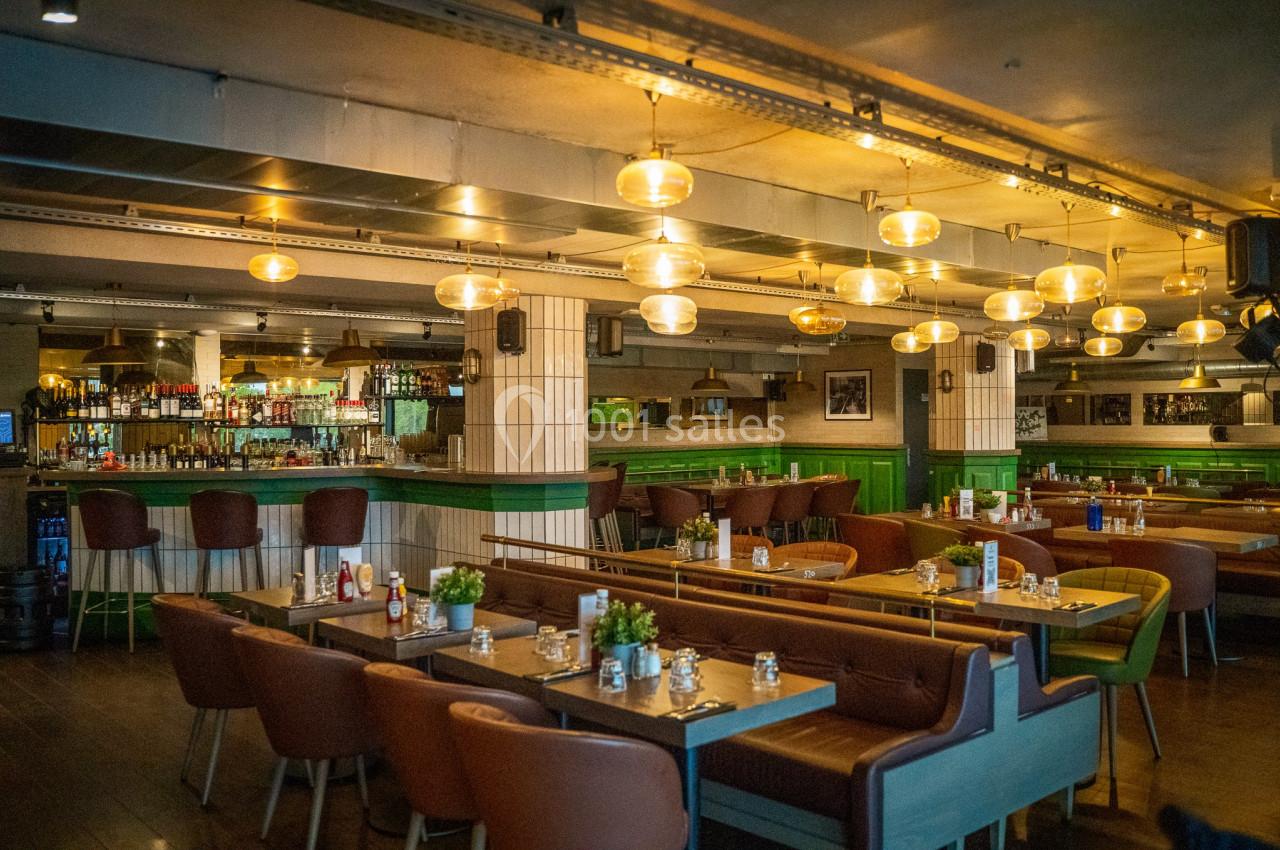 Salle de restaurant lumineuse avec des tables en bois, des banquettes, un bar en arrière-plan et des suspensions dorées.