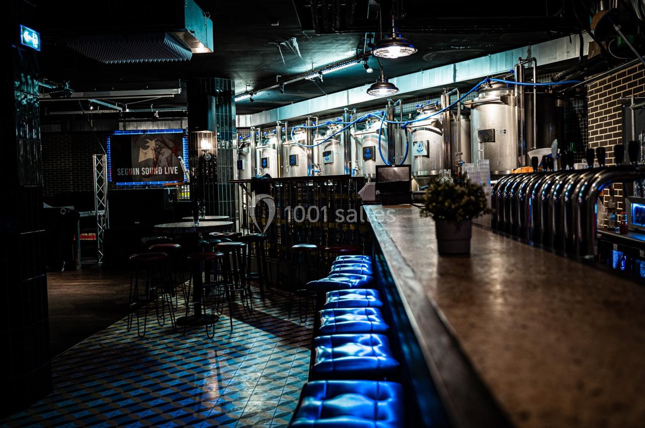 Intérieur d'un bar industriel avec des cuves en acier, un comptoir éclairé et des tabourets rembourrés bleus.
