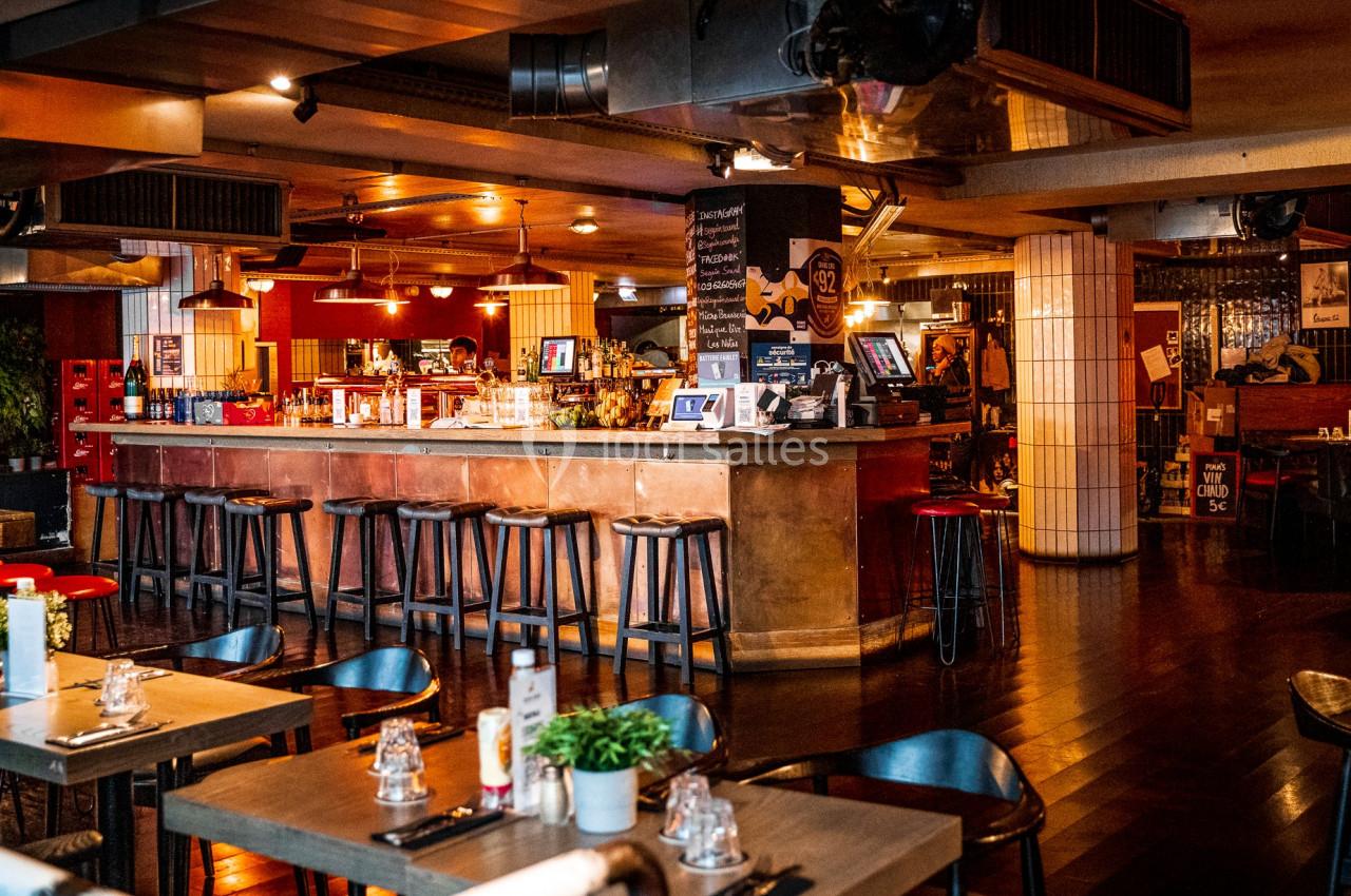 Intérieur d'un restaurant avec un bar central, des tabourets hauts, des tables dressées et une ambiance chaleureuse.