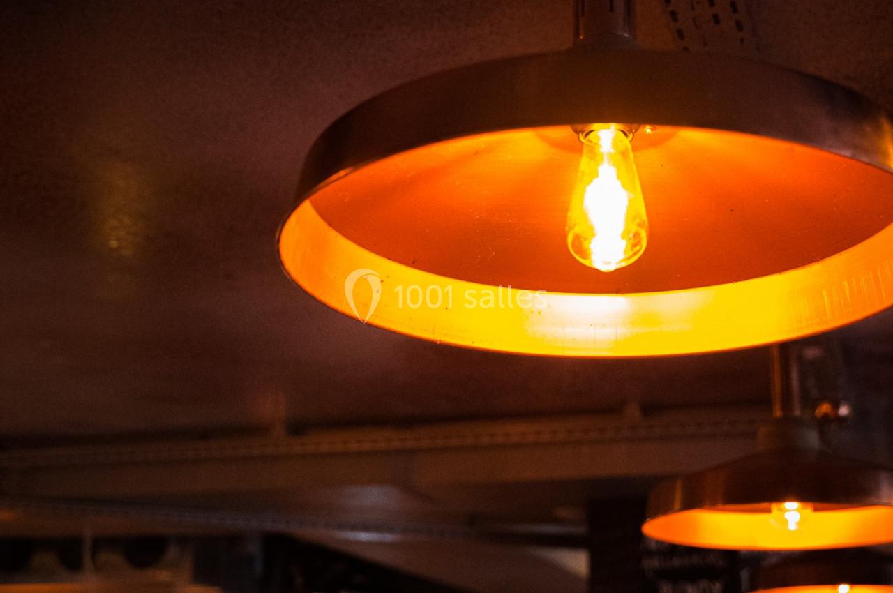 Suspension métallique orange éclairée, suspendue au plafond d'un bar avec un comptoir et des bouteilles en arrière-plan.