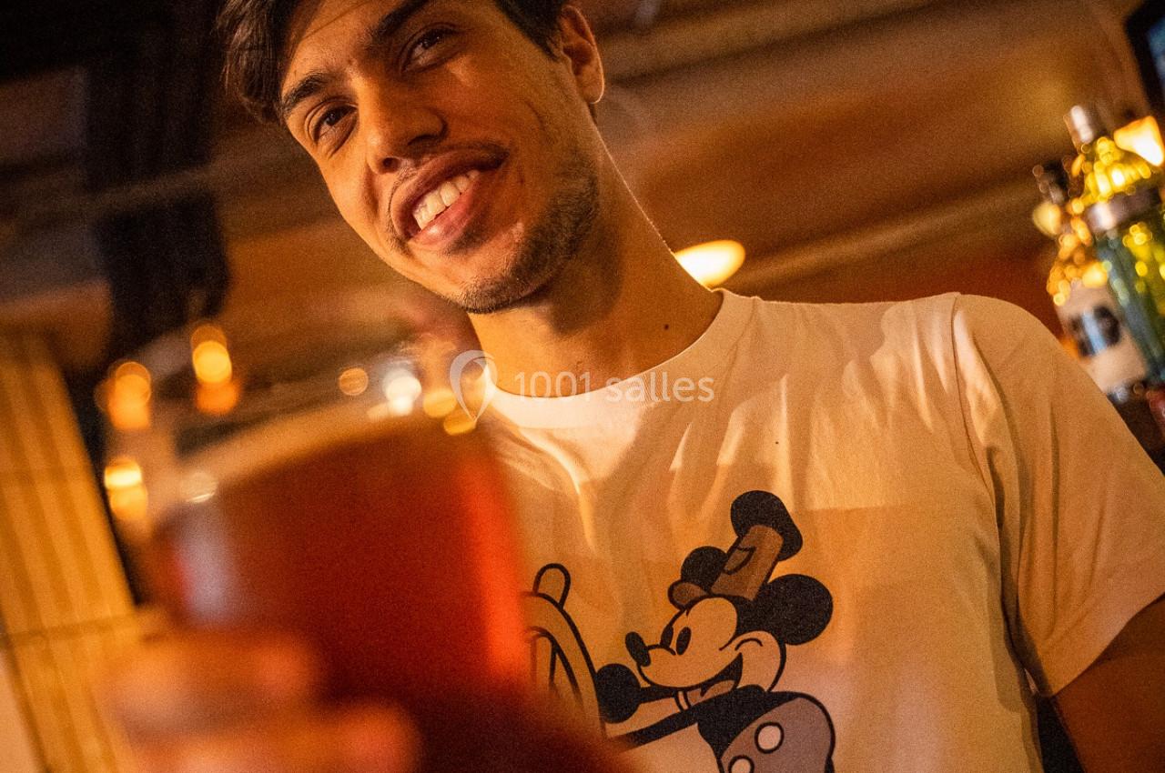 Un homme souriant tend un verre de bière dans un bar à l'éclairage chaleureux.