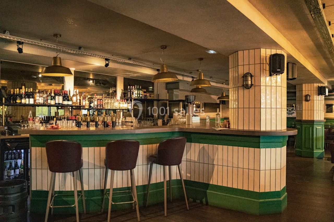 Bar avec comptoir en bois, tabourets hauts, étagères remplies de bouteilles et éclairage suspendu dans une ambiance…