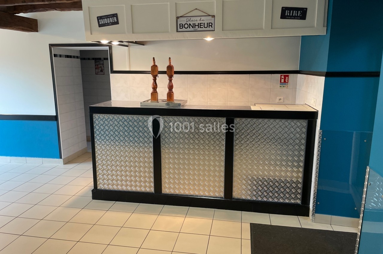 Comptoir avec deux tireuses à bière, carrelage clair, murs bleus et blancs, et poutres apparentes au plafond.