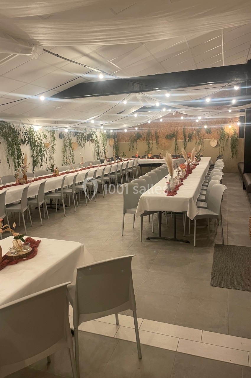 Salle décorée pour un événement, avec longues tables dressées, nappes blanches, guirlandes lumineuses et ornements végétaux.