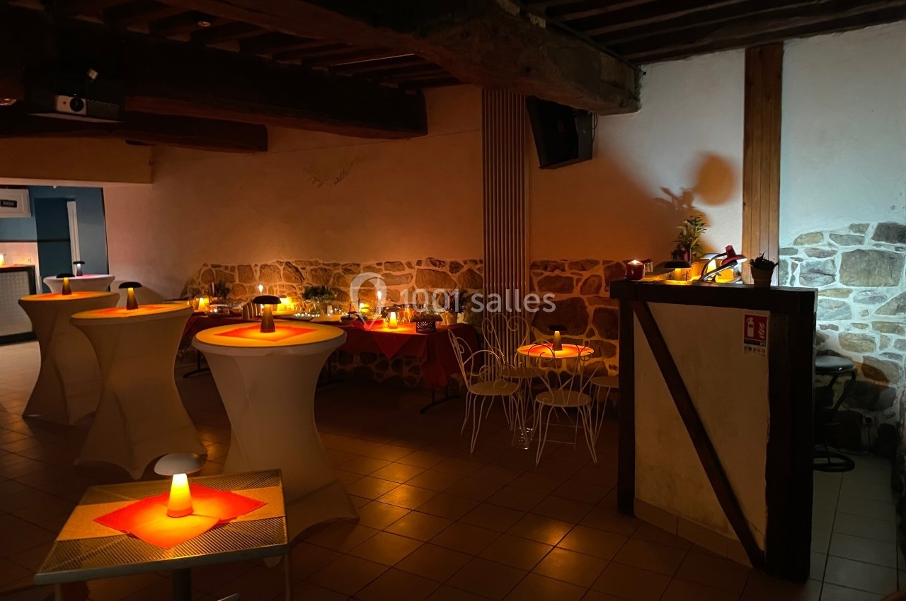 Salle faiblement éclairée avec des tables hautes et basses décorées de bougies, murs en pierre et poutres apparentes.