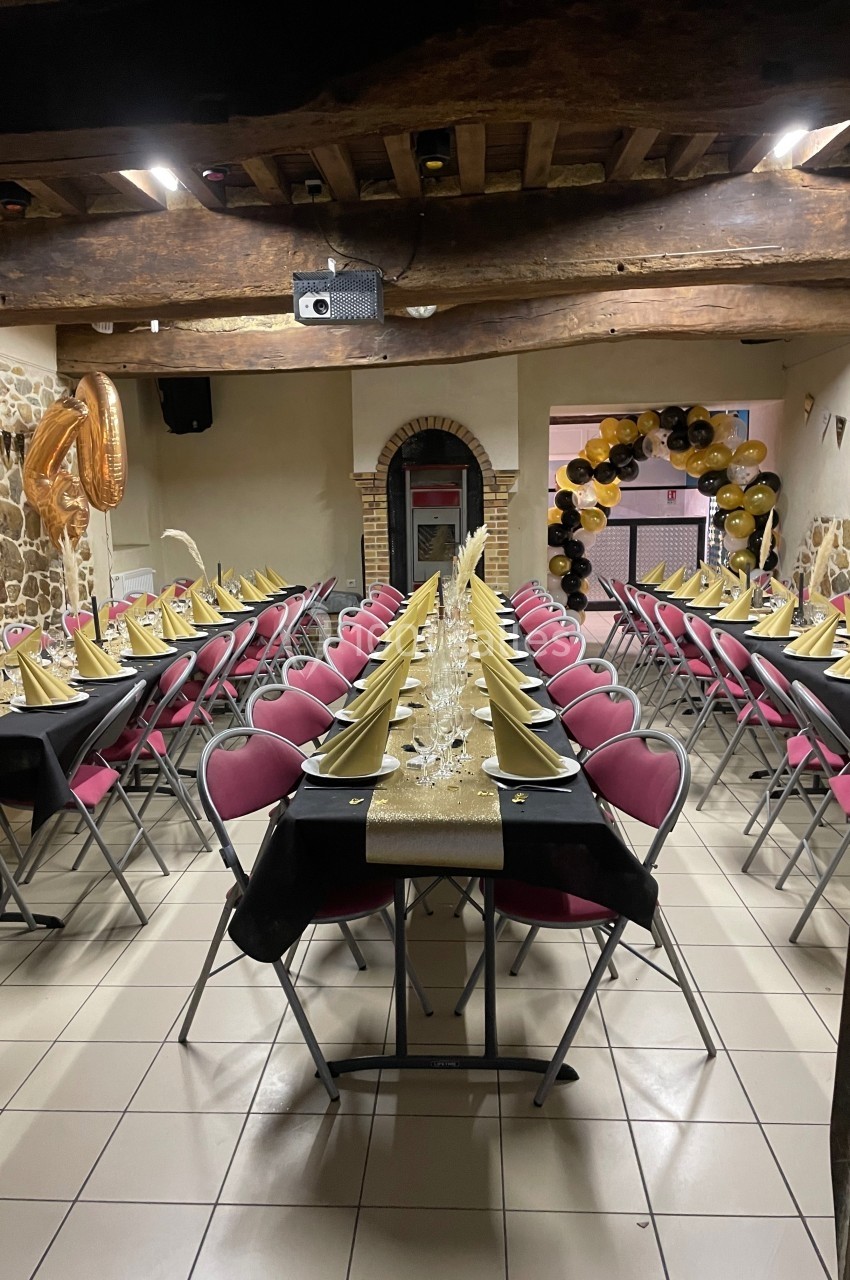 Salle décorée pour un événement avec tables alignées, nappes noires, chemins dorés, chaises roses et ballons en arrière-plan.