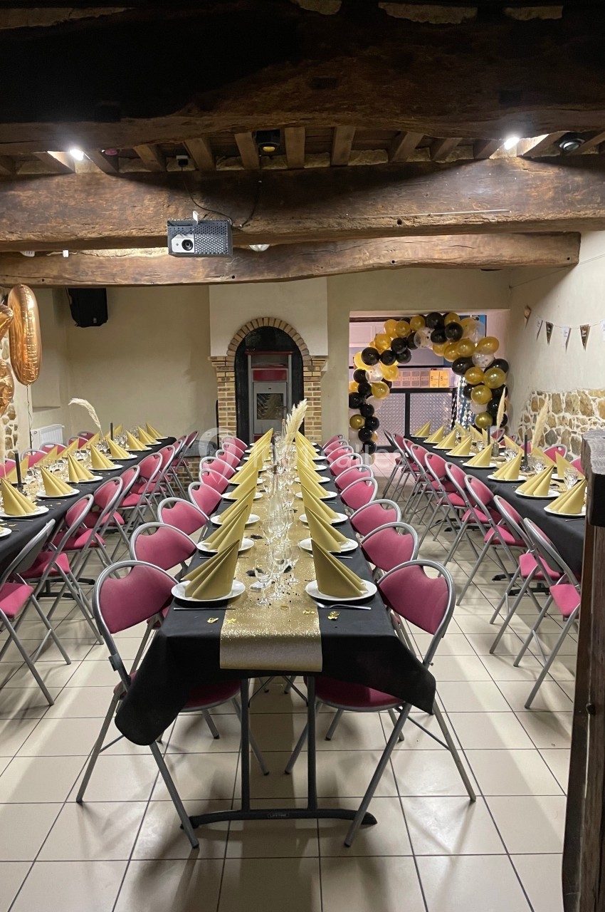 Salle décorée pour un événement, avec tables alignées, nappes noires, serviettes dorées et ballons en arrière-plan.