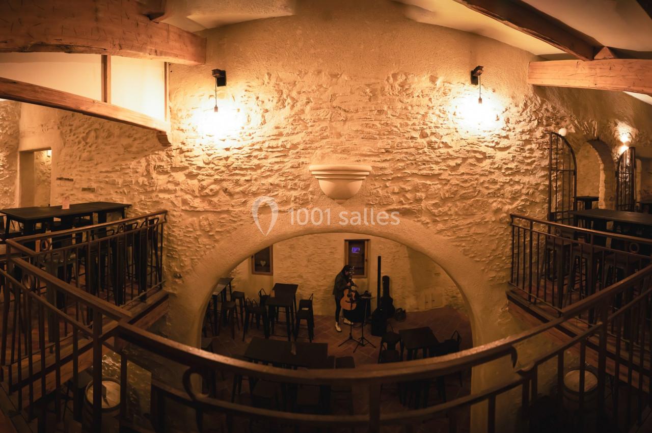 Salle intérieure en pierre avec mezzanine, éclairée par des lumières chaudes, tables et chaises disposées en bois sombre.