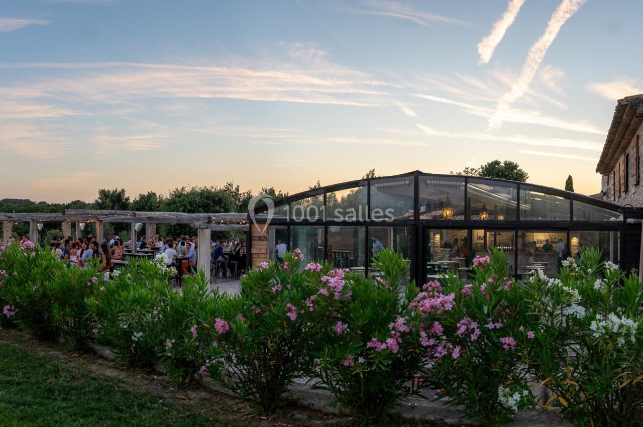 Bâtisse en pierre avec verrière, terrasse animée et jardin fleuri, sous un ciel au coucher du soleil.