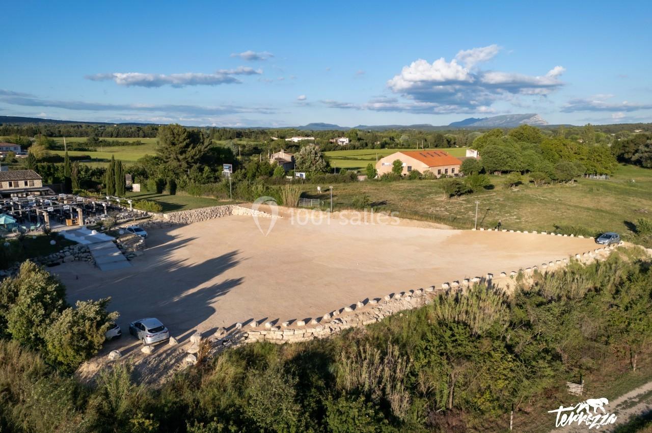Vue aérienne d'un grand parking en gravier entouré de verdure, avec des bâtiments et des collines à l'arrière-plan.