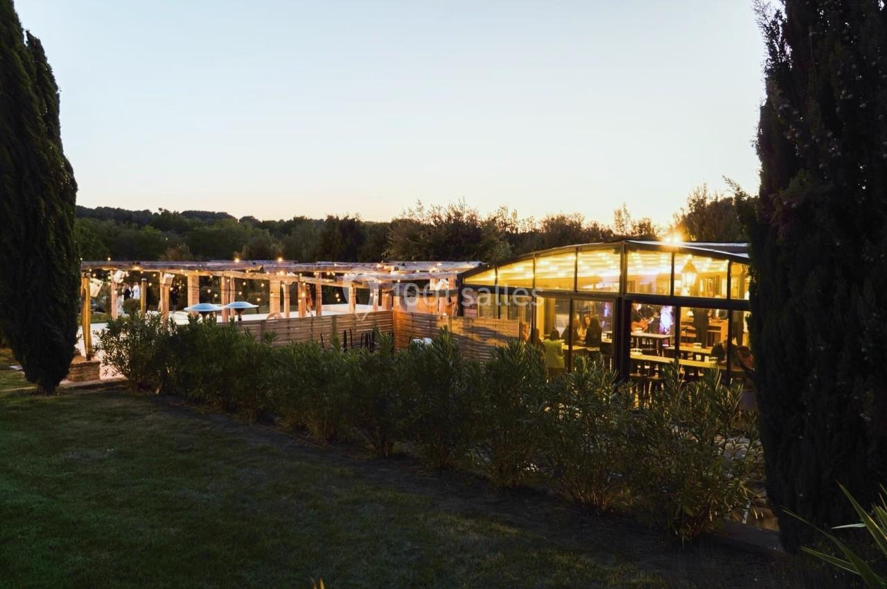 Vue d'un restaurant éclairé en soirée, entouré de végétation et d'arbres, avec une terrasse illuminée.