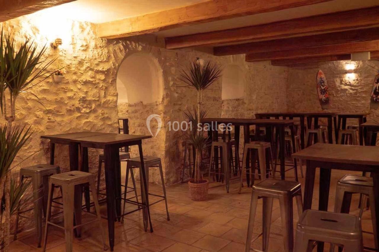 Salle avec murs en pierre, tables hautes en bois, tabourets métalliques et plantes décoratives sous un éclairage chaleureux.