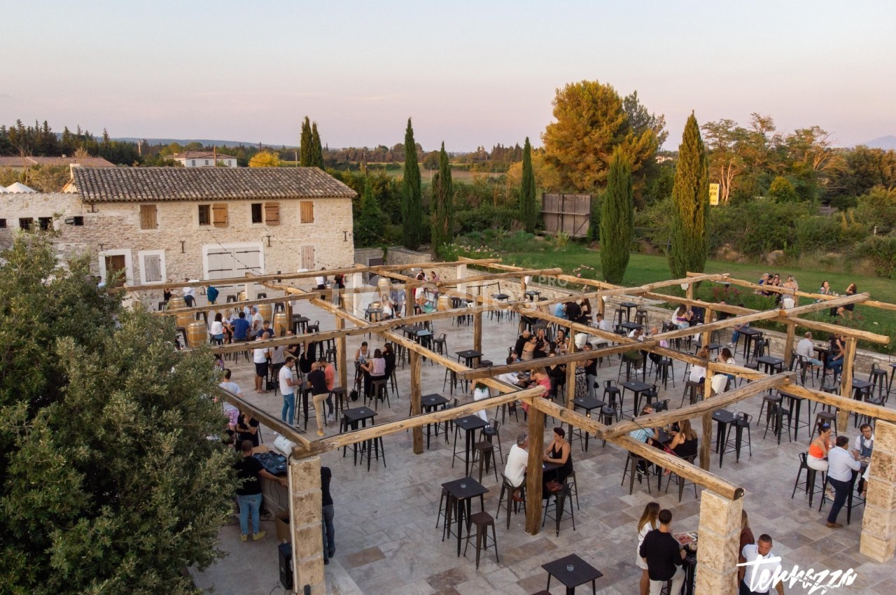 Vue aérienne d'une terrasse en pierre avec des tables hautes, des invités debout et un bâtiment en arrière-plan.