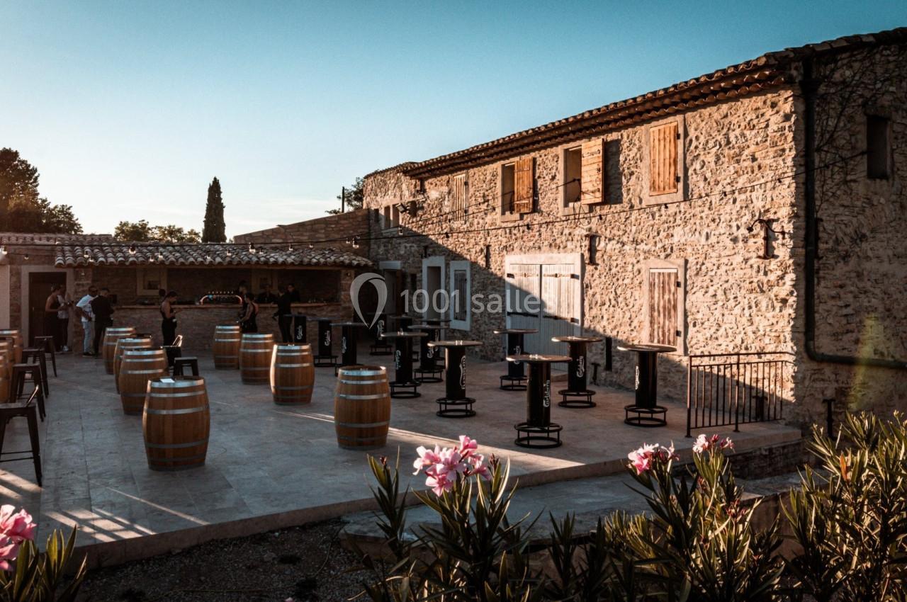 Terrasse ensoleillée avec tables, tonneaux en bois et bâtiment en pierre, entourée de végétation.