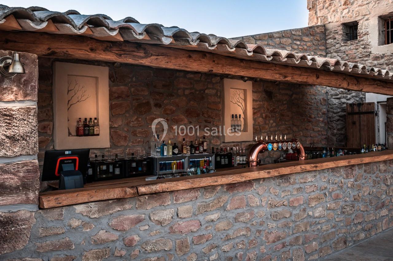 Bar extérieur en pierre avec comptoir en bois, aligné de bouteilles et de tireuses à bière sous un ciel dégagé.