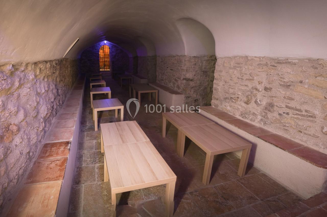 Salle voûtée en pierre avec des bancs et des tables en bois, éclairée par une lumière colorée provenant d'une fenêtre.