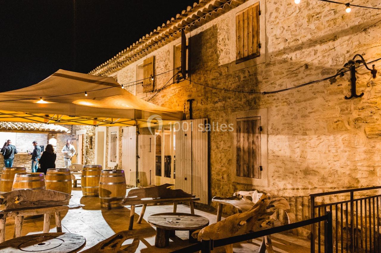 Cour extérieure éclairée d'une bâtisse en pierre, avec tonneaux, tables en bois et un auvent beige.