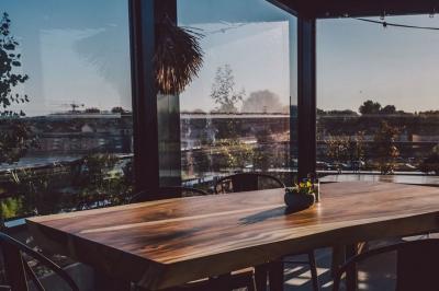 Salle lumineuse avec des tables en bois, des chaises hautes, des plantes et des stores laissant passer la lumière naturelle.