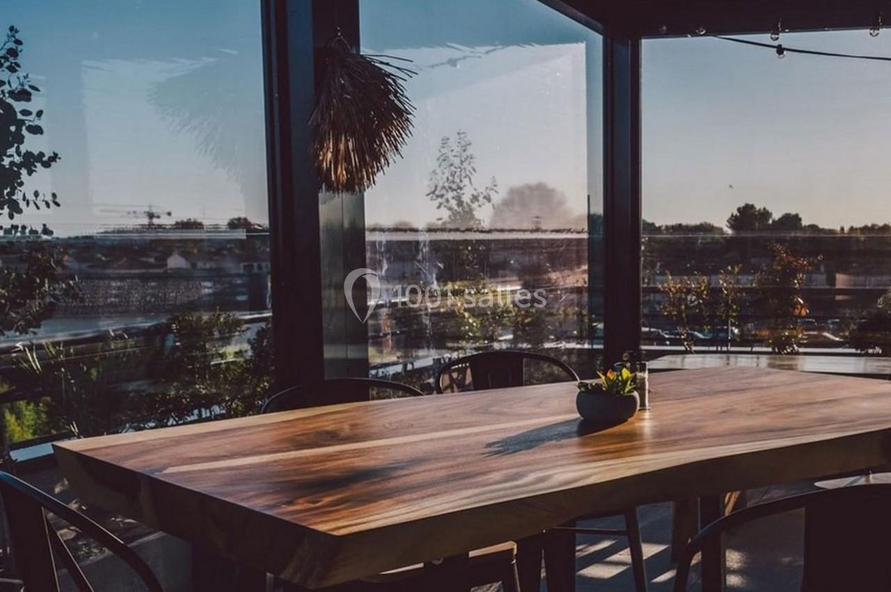 Table en bois massif entourée de chaises, placée dans un espace vitré avec vue sur un paysage extérieur ensoleillé.