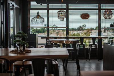 Salle lumineuse avec des tables en bois, des chaises hautes, des plantes et des stores laissant passer la lumière naturelle.