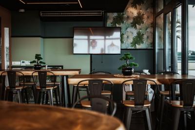 Salle lumineuse avec des tables en bois, des chaises hautes, des plantes et des stores laissant passer la lumière naturelle.