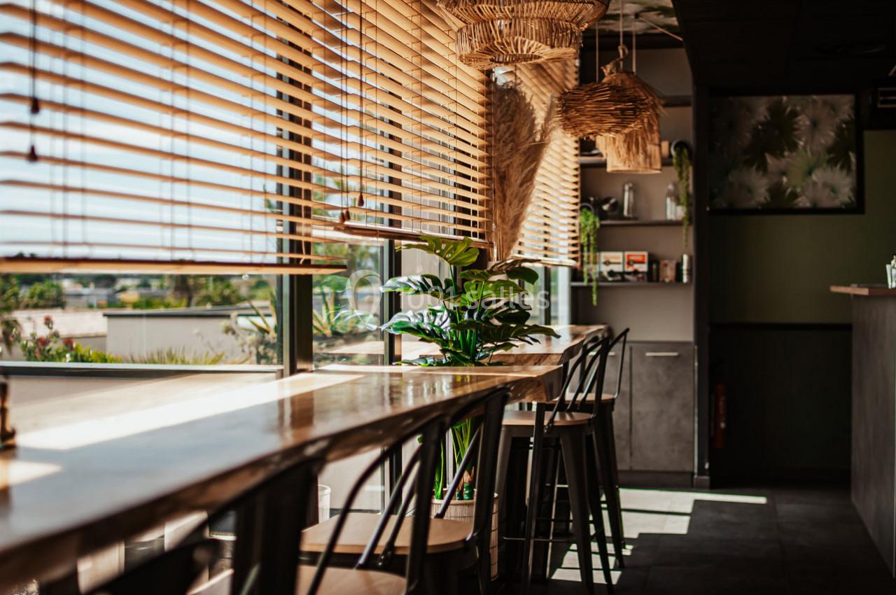 Salle lumineuse avec des tables en bois, des chaises hautes, des plantes et des stores laissant passer la lumière naturelle.