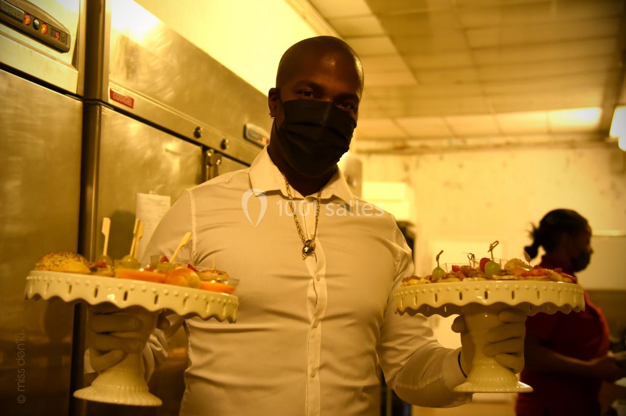 Un serveur portant un masque noir tient deux plateaux garnis de petits sandwichs et amuse-bouches dans une cuisine.