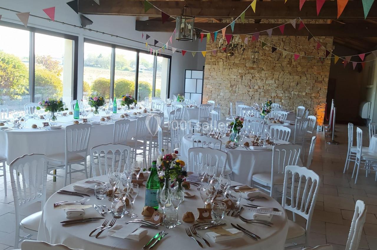 Salle de réception décorée avec des tables rondes dressées, des guirlandes colorées et une grande baie vitrée.